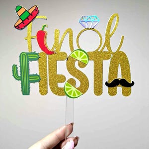May include: A gold glitter "Final Fiesta" cake topper featuring a sombrero, chilli pepper, cactus, lime slice, and moustache. The letter "l" is replaced with a diamond ring. The topper is held by a hand with a pink nail design.