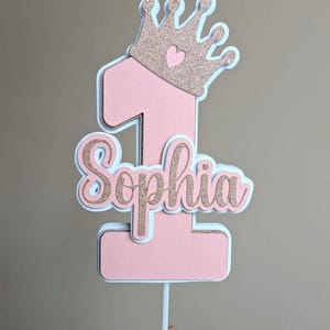 May include: Pink and gold glitter cake topper with a crown and the number one. The name Sophia is written in gold glitter below the number.