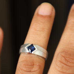 Op de afbeelding: Een zilveren ring met een blauwe vierkante edelsteen in het midden. De ring wordt aan een vinger gedragen.