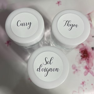 May include: Three small, clear spice jars with white lids. Each lid has a white label with black script. The labels read "Curry", "Thym", and "Sel d'oignon". The jars are arranged on a floral patterned surface.