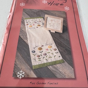 Può includere: Una confezione con un motivo floreale ricamato. Il design include un asciugamano color crema e un'immagine incorniciata, entrambi con fiori ricamati. I testi "Nutmeg Hare" e "Garden Fancies" sono visibili. La confezione è sigillata in plastica trasparente.