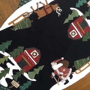 Op de afbeelding: Een zwarte kerstboomrok van vilt met een witte rand. De rok heeft een rode schuur, een rendier, een kerstman en verschillende groene bomen. De tekst "KRINGLE TREE FARM" is zichtbaar op de rok.