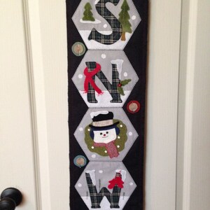May include: A black felt banner with the word "SNOW" spelled out in white felt letters. Each letter is decorated with different winter-themed elements, including trees, snowflakes, and a snowman.