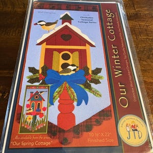 May include: A packaged wall hanging kit featuring a whimsical birdhouse design. The kit includes a red birdhouse with a blue bow, birds, and the text "Our Winter Cottage." Finished size is 10.5" x 22".