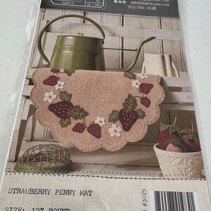Könnte beinhalten: Ein Erdbeer-Penny-Matte mit einem gewellten Rand, bestickt mit Erdbeeren und weißen Blumen. Die Matte hat einen Durchmesser von etwa 30 cm. Die Verpackung enthält eine grüne Gießkanne und eine weiße Schüssel mit Erdbeeren.