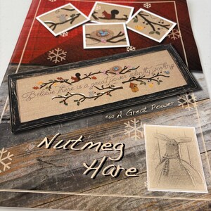 May include: A cross-stitch pattern book titled "Nutmeg Hare" featuring a framed cross-stitch design with the text "Believe There is a great power silently working." The book also shows several small cross-stitch designs of birds and squirrels.