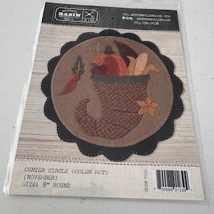 May include: A package containing a round, decorative woolen mat with a fall theme. The mat features a brown boot filled with pumpkins and leaves, surrounded by a black scalloped edge. The text on the package reads "Center Circle Woolen Mats (November) Size: 8" Round."
