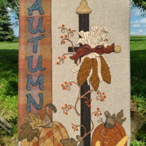 May include: A beige fabric wall hanging with the word "AUTUMN" embroidered in blue on the left side. The right side features a brown and black pole with a white and brown corn stalk, brown leaves, and orange berries. There are three pumpkins at the bottom, one green, one orange, and one yellow.