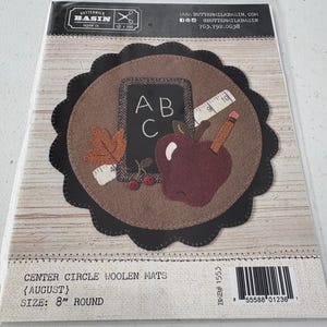 Puede incluir: Un paquete que contiene una alfombra redonda de fieltro con temática escolar. La alfombra presenta una pizarra con ABC, una manzana roja, un lápiz y una cinta métrica. La alfombra tiene 20 cm de diámetro y un borde festoneado.