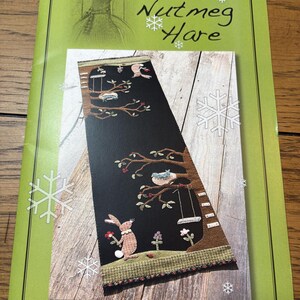 May include: A Nutmeg Hare pattern booklet with a light green cover featuring white snowflake graphics. The center displays a rectangular image of a black fabric panel with an embroidered tree, a brown rabbit, birds, and a swing, titled #51 Will you be my Friend?