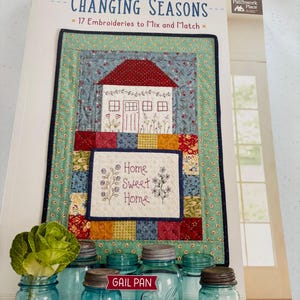 May include: A book titled "Changing Seasons" with a colorful quilt design on the cover. The quilt features a house and the words "Home Sweet Home." Several blue glass jars with metal lids are in front of the book.