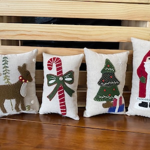 May include: Four small, rectangular, cream-colored pillows with different Christmas-themed felt appliques. The pillows are arranged in a row on a wooden surface. The appliques include a reindeer, a candy cane with a bow, a Christmas tree, and Santa Claus.