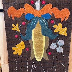 May include: A brown felt Thanksgiving banner with a stitched image of a corn stalk with a bow and leaves. The banner says "THANKS" in white thread.