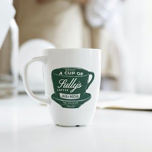 Puede incluir: Taza de café de cerámica blanca con un gráfico verde que dice "Enjoy a cup of Sully's Coffee Java & Mocha The Sullivans Chicago". La taza tiene un asa curva y está sobre un fondo borroso.