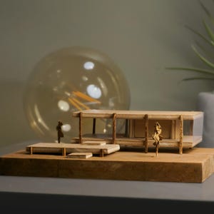 May include: A wooden model of a modern house with a glass wall and a flat roof. The model is on a wooden base and includes small figures of people.