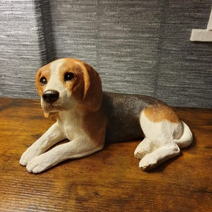 May include: A beagle dog figurine in a lying position, featuring a tri-colour coat of brown, black, and white. The dog has long, floppy ears, a white muzzle, and a black nose, with its front paws extended forward and hind legs tucked.