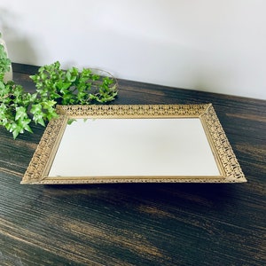 May include: A gold-colored rectangular tray with a decorative, ornate frame and a mirrored surface.