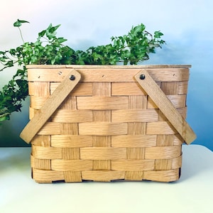 May include: A rectangular woven wooden basket with a light tan finish. The basket features a flat lid and two hinged wooden handles attached with metal rivets on each side. The construction consists of wide, horizontal and vertical strips.