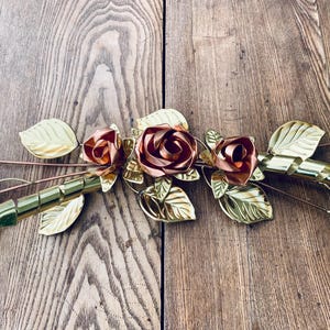 May include: Metal rose garland with copper and gold-colored roses and leaves. The roses are a deep copper color, and the leaves are a bright gold. The garland is arranged on a wooden surface.