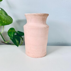 May include: A peach-colored, handcrafted vase with a textured surface. The vase has a wide base, a narrow neck, and a slightly flared rim. The top of the vase has horizontal ridges. Green leaves and vines are visible in the background.