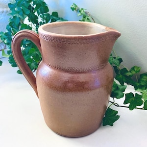 May include: A brown ceramic pitcher with a rounded body, a curved handle, and a wide pouring spout. The upper section of the pitcher features a textured band with a repeating pattern, while the interior is a smooth, light cream color.
