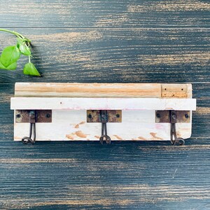 May include: A rustic, white-painted wooden wall hook with three metal hooks. The wall hook is mounted on a dark blue wooden surface. A small green plant is in the upper left corner.
