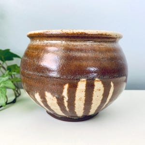 May include: A handcrafted ceramic pot with a rounded shape. The pot features a rich brown glaze with lighter beige vertical stripes near the base. The rim is a lighter tan color. A decorative piece for the home.