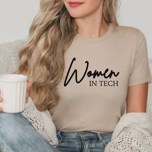 May include: A woman wearing a beige t-shirt with the text "Women in Tech" in black script. She is sitting on a couch and holding a white mug.