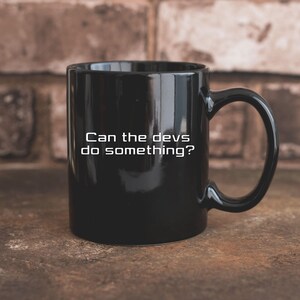 Puede incluir: Taza de cerámica negra con asa curva. La taza presenta el texto blanco: "Can the devs do something?" La taza está colocada contra una pared de ladrillos y una superficie marrón.