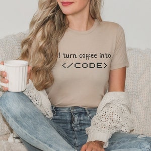May include: A woman wearing a beige t-shirt with the text "I turn coffee into <CODE>" printed on it. She is holding a white mug and sitting on a white couch.