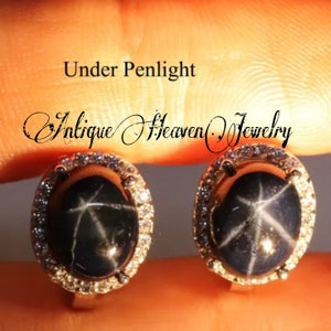 May include: A pair of silver earrings with a black star-shaped gemstone surrounded by a halo of smaller white gemstones. The earrings are shown under a penlight, highlighting the sparkle of the gemstones. "Under Penlight" and "Antique Heaven Jewelry" are written above the earrings.