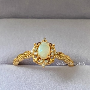 Puede incluir: Un anillo dorado con una piedra preciosa de ópalo ovalada en el centro, rodeada de pequeñas piedras preciosas transparentes. El anillo tiene un diseño ornamentado de inspiración vintage con detalles delicados. El texto "Antique Heaven Jewelry" es visible.
