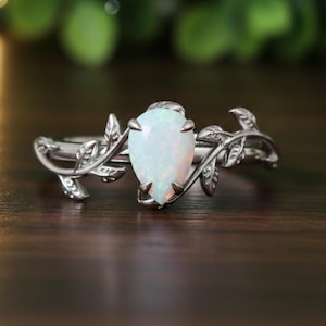 May include: A silver ring featuring a pear-shaped, white opal gemstone. The ring's band is adorned with delicate leaf and vine detailing, creating an elegant, nature-inspired design. The opal is set in a prong setting.
