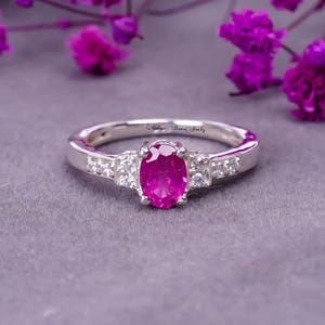 Peut inclure: Une bague en argent avec une pierre ovale rose entourée de petites pierres blanches. La bague est posée sur un fond gris.
