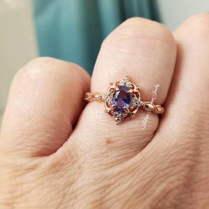 Könnte beinhalten: Roségoldener Ring mit einem ovalen, tiefvioletten Edelstein, umgeben von kleinen weißen Steinen. Der Ring hat ein verziertes, vintage-inspiriertes Design mit filigranen Details am Band. Der Text "Antique Queen Jewelry" ist sichtbar.