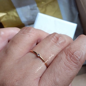 Puede incluir: Un delicado anillo de oro con una pequeña gema redonda de color rosa claro. El anillo se lleva en un dedo. La imagen también muestra una caja blanca y una superficie dorada.