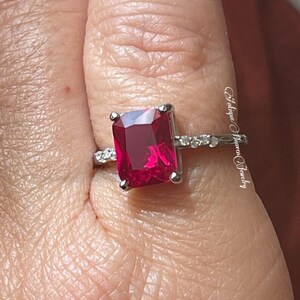 Pu&ograve; includere: Un anello d'argento con una grande pietra preziosa rossa quadrata brillante circondata da piccole pietre preziose chiare. L'anello &egrave; su un dito.