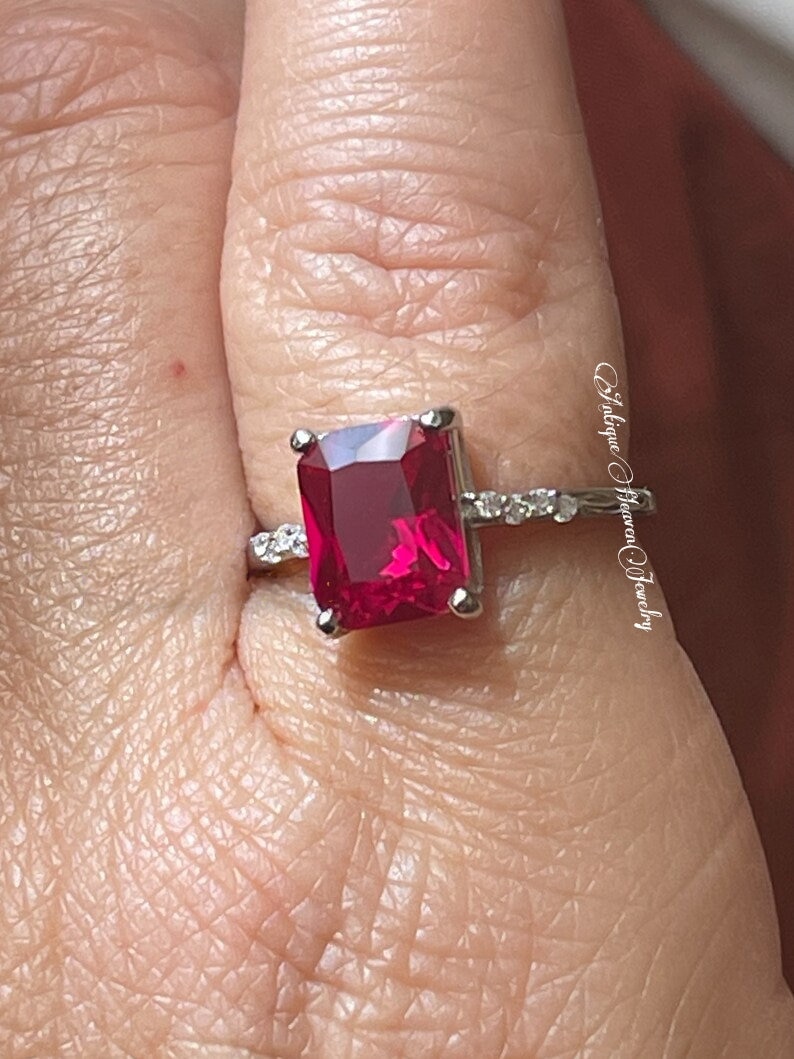 Pu&ograve; includere: Un anello d'argento con una grande pietra preziosa rossa quadrata brillante circondata da piccole pietre preziose chiare. L'anello &egrave; su un dito.