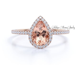 May include: A pear-shaped engagement ring with a halo setting. The center stone is a peach-colored gemstone surrounded by a row of smaller, clear gemstones. The band is rose gold and has a row of clear gemstones.