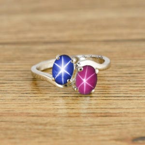 May include: A silver ring featuring two oval-shaped gemstones. The left stone is a blue star sapphire and the right stone is a pink star ruby.