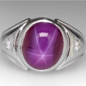 May include: A silver ring with a large, oval-shaped, purple star sapphire gemstone. The gemstone has a six-pointed star in the center. The ring has two small diamonds on either side of the main stone. "Antique Hawaiian Jewelry" is inscribed on the band.