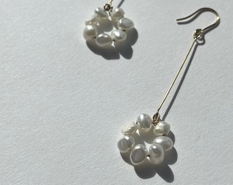 Può includere: Un paio di orecchini pendenti color oro. Ogni orecchino presenta un gruppo circolare di perle bianche sospese a un lungo e sottile filo d'oro. Gli orecchini sono esposti su una superficie bianca, con ombre.