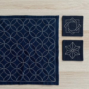 Puede incluir: Tres posavasos de tela azul oscuro con costuras blancas. El posavasos más grande tiene un patrón repetitivo de círculos y los dos posavasos más pequeños tienen diferentes diseños geométricos.