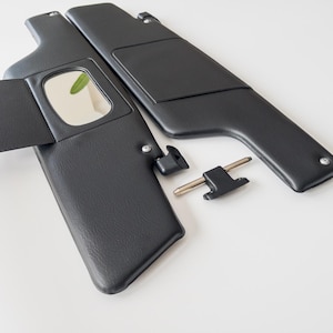 May include: Two black car sun visors with a mirror and a small, attached flap. The visors are made of a textured material and are accompanied by mounting hardware. The items are set against a white background.