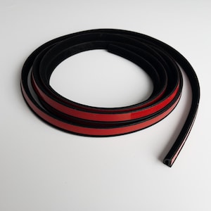 Puede incluir: Una tira de goma negra enrollada con un respaldo adhesivo rojo. La tira es flexible y parece ser para sellar o bordear. La imagen está tomada sobre un fondo blanco.