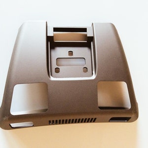 Può includere: Una custodia in plastica marrone con un'apertura rettangolare e due aperture più piccole sui lati. La custodia ha una superficie testurizzata e una piccola ventilazione sul fondo.