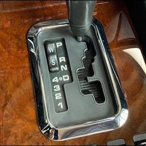 May include: An automatic transmission gear shifter console featuring a black gate pattern with white lettering for P, R, N, D, 4, 3, 2, 1, and W/S modes. The shifter assembly is framed by a polished chrome border and set into a glossy wood grain panel.