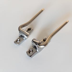 May include: Two chrome-plated metal hinges with a curved lever and mounting plate. The levers have a gold-coloured tip. The hinges are designed for a door or cabinet and are shown on a white background.