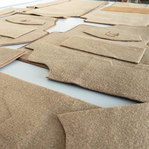 May include: A collection of tan car floor mats, cut and arranged on a light surface. The mats vary in size and shape, with some featuring curved edges and cutouts. The material appears to be a soft, textured fabric, possibly carpet or felt.