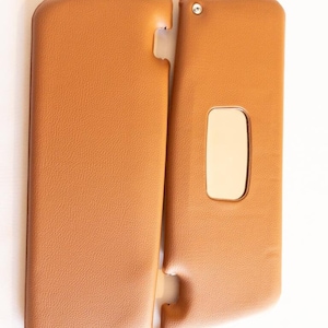 May include: A pair of brown leather sun visors with a small mirror on one visor. The visors are attached together with a plastic clip.